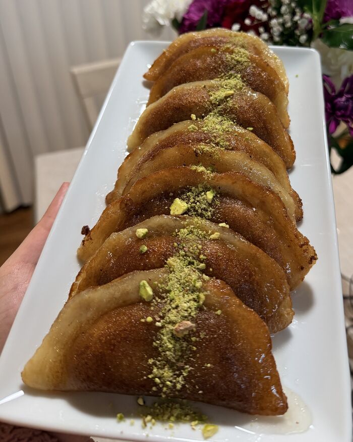 A long white plate of atayef, folded and pan-fried to a deep golden brown, glossy with syrup and dusted down the middle with finely ground pistachio.